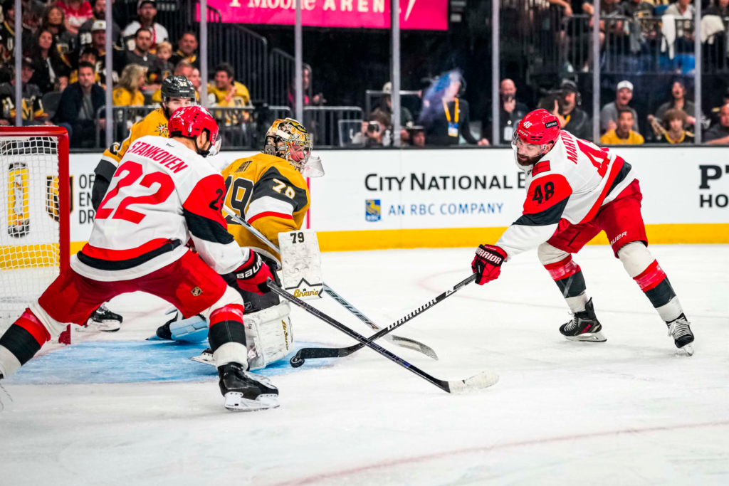 Jogador do Carolina disputa o disco com o goleiro de Vegas, no jogo 3 da final da Stanley Cup 2026, entre as equipes de Vegas Golden Knights e Carolina Hurricanes - Foto: © Josh Lavallee/Carolina Hurricanes – Fonte: NHL (2026)